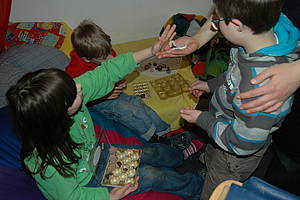 Drei Schüler sitzen auf dem Bett, zwischen ihnen liegen Pralinenpackungen, ein Mädchen reicht einer anderen Person "Schokolade"