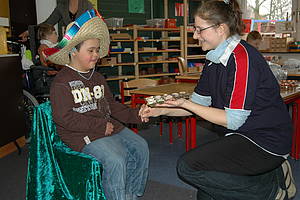 Ein Schüler sitzt auf einem Stuhl und bekommt von der Erzieherin Schokolade überreicht