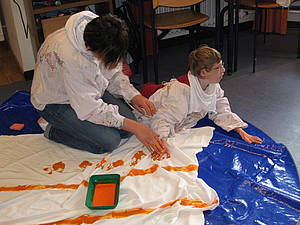 Eine Schülerin liegt auf dem Boden und verziert mit Hilfe ihrer Einzelfallhelferin ihren Stoff mit Handabdrücken.