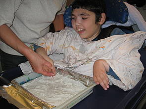 Ein Schüler sitzt lachend im Rollstuhl, während eine Mitarbeiterin seine Hand durch eine Schale mit Gips führt.
