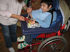 Ein Schüler sitzt im Rollstuhl, während eine Mitarbeiterin ein vor ihm liegendes Gipsrelief mit ihm gemeinsam bemalt.
