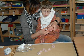 Eine Schülerin sitzt auf dem Schoß ihrer Einzelfallhelferin und wickelt mit ihr Gips-Schokolade in Alu-Folie ein